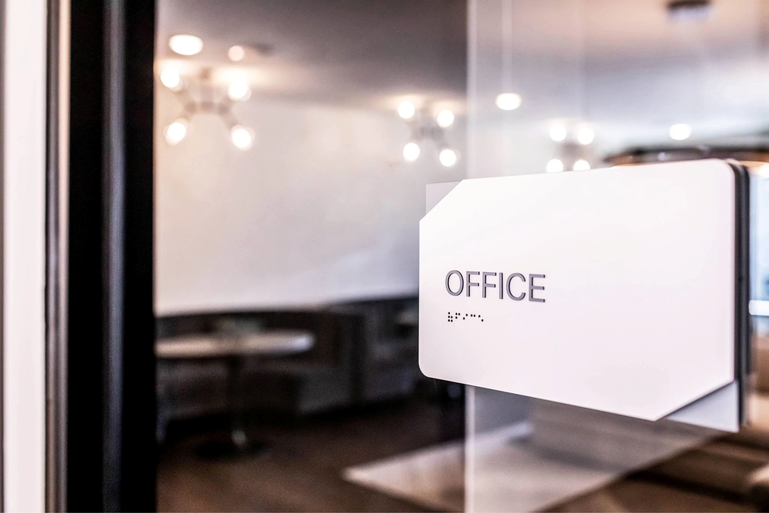 A sleek glass door featuring a white office sign with braille lettering leading into a modern apartment workspace.